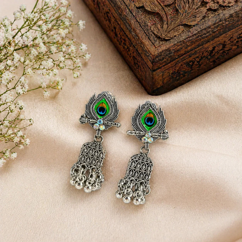 Peacock eye jhumka earrings with green enamel, silver filigree, and dangling bells on soft fabric