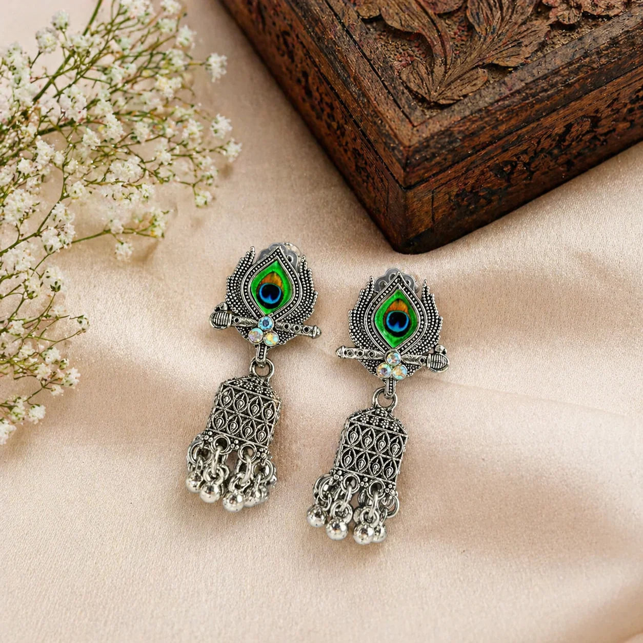 Peacock eye jhumka earrings with green enamel, silver filigree, and dangling bells on soft fabric