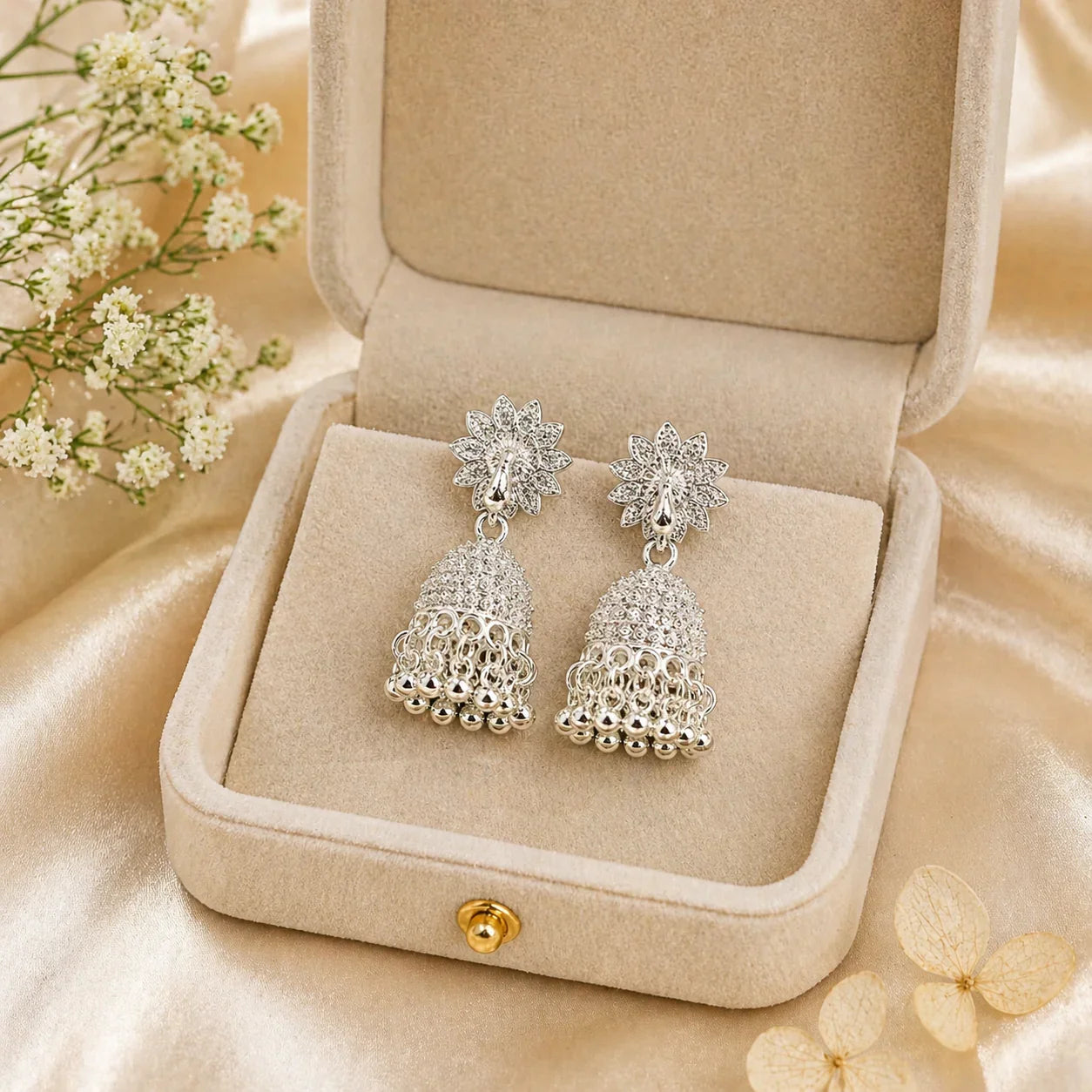 Elegant silver jhumka earrings with floral studs, displayed in a velvet box on satin fabric with delicate flowers.