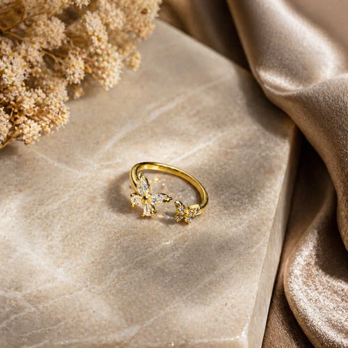 Gold butterfly ring with clear gemstones on marble, dried flowers, and satin fabric